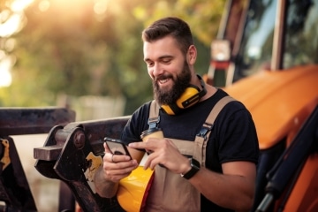 Young architect leaning up against construction machinery, smiling at phone and drinking coffee to go