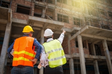 Two men wearing hi-vis vests and hard hats looking up at a construction site, the one on the right is pointing up at the site