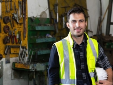 Smiling man wearing yellow hi-vis vest holding a white hard hat with a wall of tools behind him