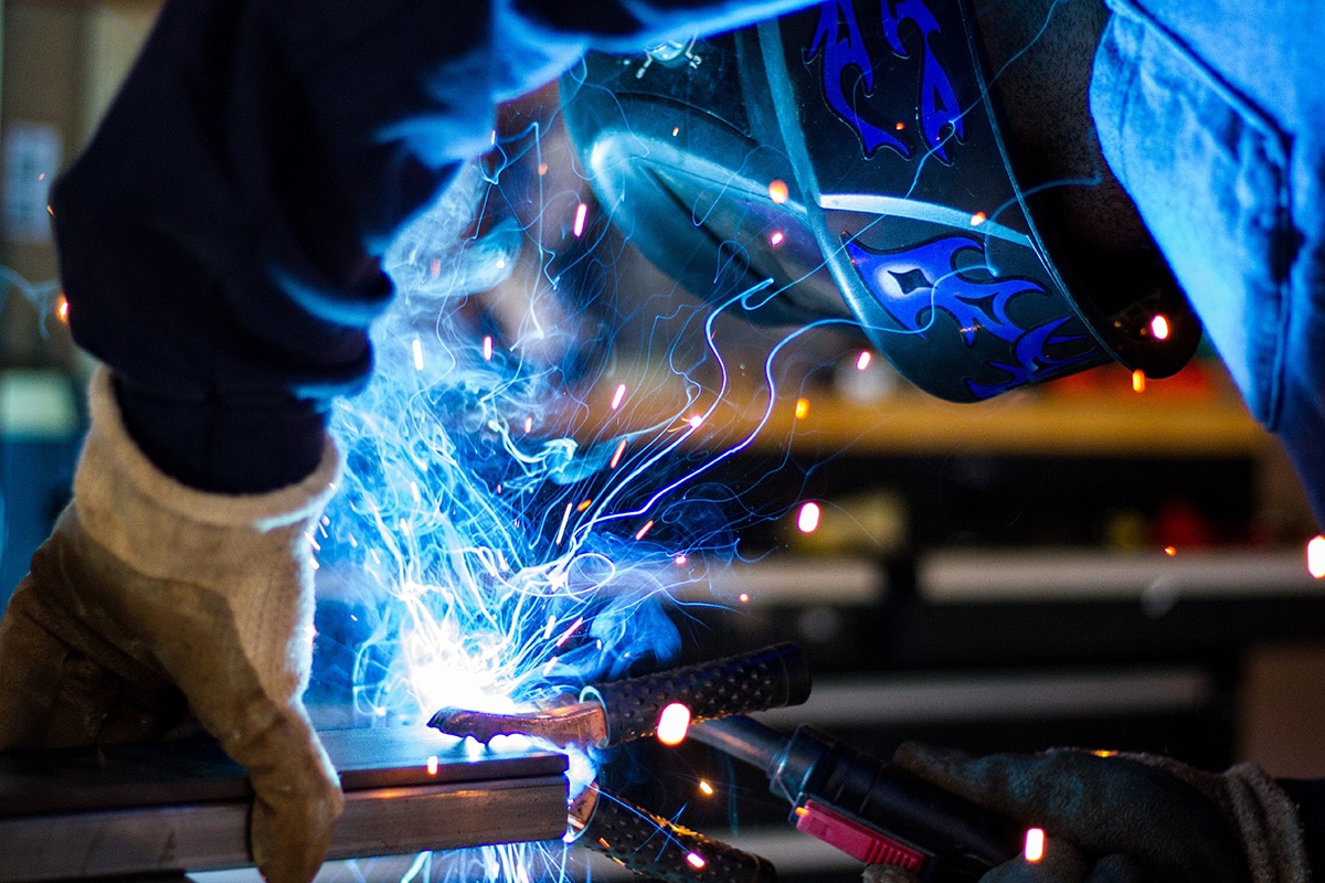 Welder wearing personal protective equipment while welding metal, blue sparks and smoke are coming from their tools that they are holding