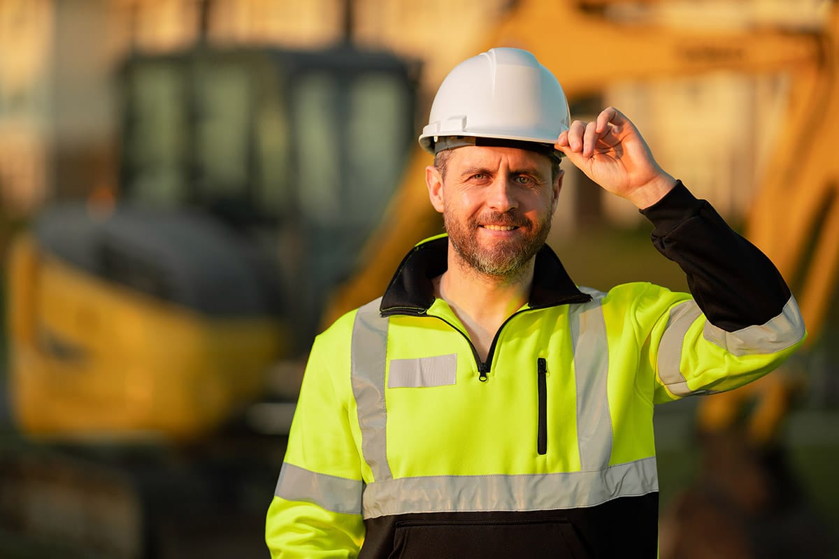 Man wearing yellow hi-vis jumper with his hand holding the front of his white hard hat, the background is blurred but behind him is earthmoving equipment